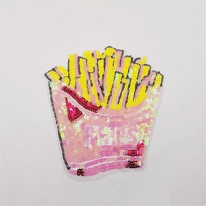 Puede incluir: Un parche de lentejuelas con forma de caja de patatas fritas. Las patatas fritas son amarillas y el recipiente es rosa con la palabra "FRIES" escrita con lentejuelas blancas. El parche está delineado en negro.