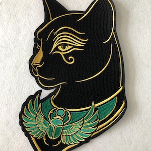 May include: Black embroidered patch featuring the profile of an Egyptian cat with gold accents. The cat has an eye of Horus design and a green and gold scarab beetle collar. The patch is likely for clothing or accessories.