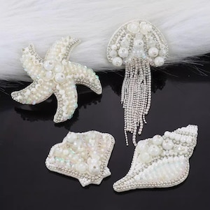 Peut inclure: Quatre patchs thermocollants en forme d'étoile de mer, de coquillage et de méduse, ornés de perles et de strass blancs.