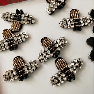 May include: A set of ten gold and black bee-shaped embellishments with clear rhinestones. The bees are made of fabric and have a 3D effect.