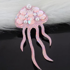 May include: A pink jellyfish-shaped applique with iridescent sequins and pearl beads. The applique has long, flowing tentacles and is designed for embellishment on clothing or accessories. The design is on a white fluffy background.