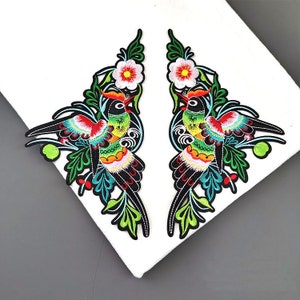 May include: Two colourful embroidered iron-on patches featuring birds with floral designs. The birds are black, green, yellow, and red with blue accents. The flowers are white with yellow centres.
