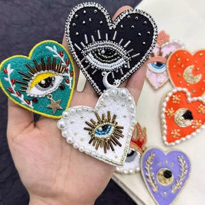 May include: Assortment of embroidered heart-shaped patches with an evil eye design. The patches are in various colours, including black, green, white, pink, orange, and purple. Each patch features a different evil eye design with beads and sequins.