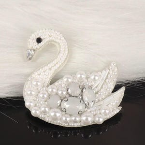 May include: A white swan brooch, embellished with pearls and crystals. The swan's body is covered in small, round pearls, with larger pearls and crystal accents on the wings and body. The swan has a black eye and a crystal beak.