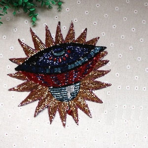 Op de afbeelding: Een gouden, rode en blauwe pailletten ooglap met een zonnestralenontwerp. De ooglap is een decoratieve versiering voor kleding of accessoires.