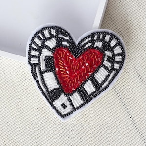 May include: A heart-shaped beaded patch with a black and white geometric pattern and a red beaded heart in the center.
