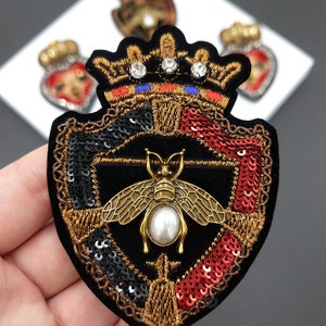 Può includere: Una toppa ricamata in oro con sfondo in velluto nero. La toppa presenta una corona con strass, uno scudo con paillettes rosse e nere e un'ape dorata con una perla.