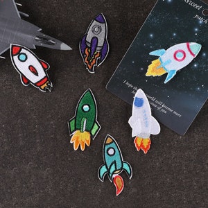 May include: Set of six colourful embroidered iron-on patches featuring cartoon rockets with flames. The patches are on a dark grey surface.  "Sweet patch" is written on a white card with a black starry background.