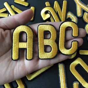 May include: Embroidered gold fabric letters spelling "ABC" with a black border. Additional gold letters are scattered in the background. These iron-on patches are ideal for customizing apparel, bags, or other fabric items.