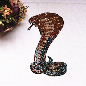 Può includere: Una toppa a forma di cobra con paillettes nei toni del marrone, oro e verde acqua. Il serpente è raffigurato con il cappuccio sollevato e la lingua estesa. La toppa è progettata per essere applicata su abbigliamento o accessori.