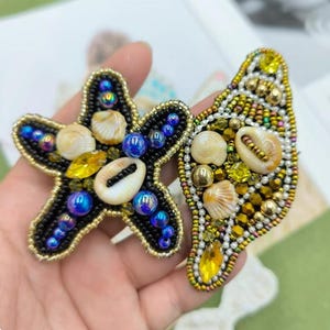 Könnte beinhalten: Zwei handgefertigte, mit Perlen besetzte Broschen. Eine in Form eines Seesterns, die andere in Form einer Muschel. Beide sind schwarz mit goldenen Verzierungen, schillernden blauen Perlen und kleinen Muscheln. Die Seesternbrosche hat gelbe und weiße Akzente.