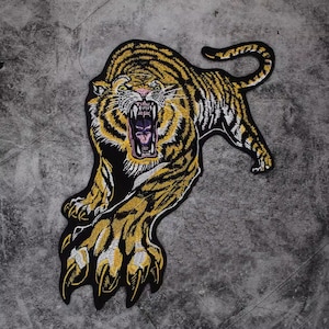May include: Embroidered tiger patch in yellow, black, and white. The tiger is depicted mid-roar with its mouth open, showcasing sharp teeth. The patch has a detailed design, with the tiger's stripes and fur clearly visible. The patch is likely for clothing or accessories.