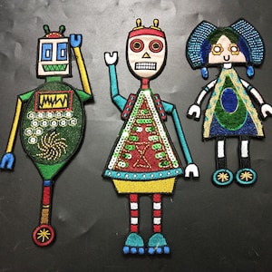 Op de afbeelding: Drie geborduurde strijkpatches met een robot, een skelet in een kleurrijk jurkje en een meisje met blauw haar en een groene jurk. De robot is groen en geel met een rood en wit wiel. Het skelet draagt een rood, geel en groen jurkje met een rood en witte schedel. Het meisje draagt een blauwe jurk met groene en blauwe accenten.