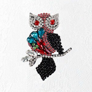 May include: A jeweled owl brooch with a black body and a branch. The owl's head is silver with red and clear crystal accents. The wings are teal, red, and green. The owl is perched on a branch with clear crystal leaves.