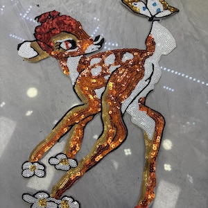 Op de afbeelding: Een met pailletten bewerkte opstrijkbare patch van een jong hert met oranje, bruine en witte vacht, voorzien van een vlinder op de staart en witte bloemaccenten bij de voeten. Het ontwerp is gemaakt met glinsterende pailletten in detail.