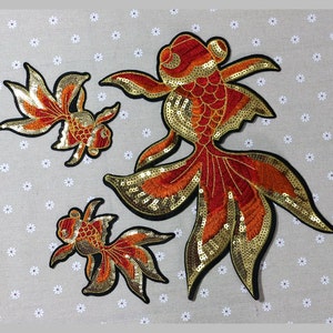 May include: Three embroidered goldfish patches in varying sizes. The fish feature orange and gold sequins, with black outlines. The patches are designed with detailed fins and tails, perfect for adding a decorative touch to clothing or accessories.