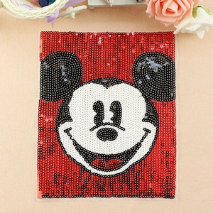 Puede incluir: Un parche de lentejuelas rojo con la cara sonriente de Mickey Mouse. La cara es blanca con detalles negros, y las orejas son negras. El fondo es rojo, creando un contraste llamativo.