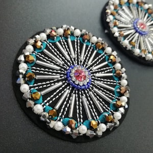 Puede incluir: Dos parches redondos de fieltro negro con un diseño circular de cuentas plateadas, blancas y doradas, lentejuelas y bordados. El centro de cada parche presenta un círculo más pequeño de cuentas azules, rosas y blancas.