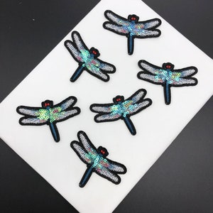 May include: Six iridescent blue dragonfly iron-on patches with red eyes. The dragonflies are made of sequins and have a white outline.