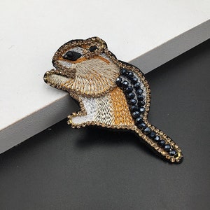 Pode incluir: Aplique bordado de esquilo com cauda de contas pretas. O esquilo é castanho e branco com detalhes em fio dourado.