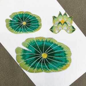 May include: Embroidered patches of a lotus flower and lily pads. The lily pads are green with yellow and white detailing, radiating from a central yellow circle. The lotus flower is green and yellow with white detailing.