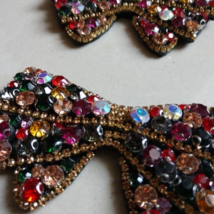 May include: Two bow-shaped embellishments, adorned with colourful rhinestones. The bows have a black base with a gold border. The rhinestones are in shades of red, pink, green, yellow, and clear, creating a sparkling effect.