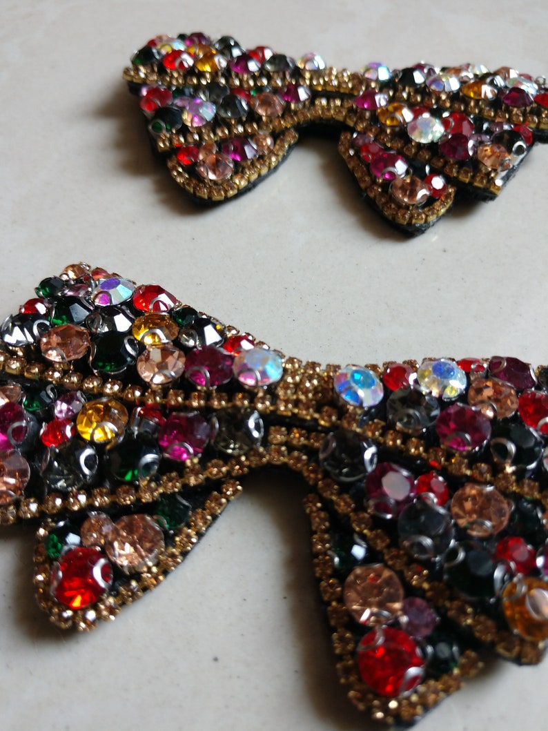 May include: Two bow-shaped embellishments, adorned with colourful rhinestones. The bows have a black base with a gold border. The rhinestones are in shades of red, pink, green, yellow, and clear, creating a sparkling effect.