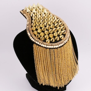 Puede incluir: Charretera dorada con base negra. La charretera presenta filas de púas doradas, un borde de cadena y largas borlas de cadena dorada. El accesorio se exhibe sobre un maniquí negro.