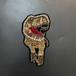 May include: A gold and brown sequined dinosaur applique with a red mouth and white rhinestones around the eyes. The applique is on a black background.