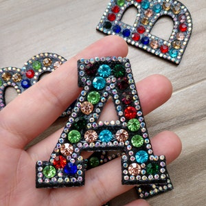 May include: A close-up of a black felt letter A covered in colorful rhinestones. The letter is being held in a hand.