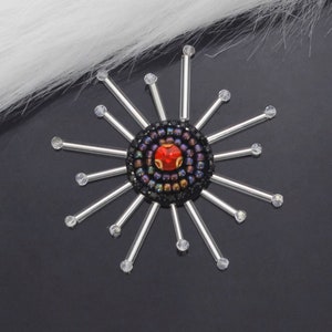 May include: A silver starburst with a red and black beaded center. The center has a red jewel in the middle.