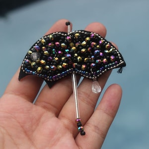 Puede incluir: Broche de paraguas negro con cuentas iridiscentes y una gota de cristal transparente. La tela del paraguas es negra y el mango es un tubo transparente. El broche se sostiene en una mano sobre un fondo azul.