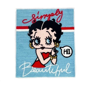 Può includere: Un toppa di tessuto blu con un'immagine di cartone animato di Betty Boop. Indossa un vestito rosso e ha un fumetto che dice "Ciao". Il testo "semplicemente bella" è scritto in corsivo bianco.