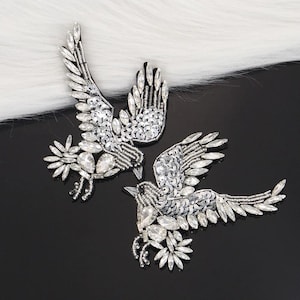 Peut inclure: Deux ornements argentés en forme d'oiseaux aux ailes déployées. Les oiseaux sont ornés de paillettes et de cristaux clairs et facettés. Les ailes et les corps sont ornés de broderies détaillées, créant un effet scintillant. Le fond est noir et blanc.