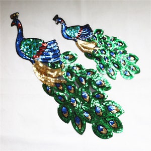 May include: Two sequined peacock appliques. The peacocks feature blue, green, and gold sequins. The peacock's body is blue and green, with a gold tail. The tail feathers are green with blue and gold accents. The appliques are on a white background.