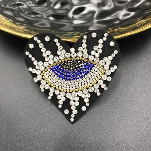 May include: A black heart-shaped patch with a blue evil eye design. The eye is surrounded by clear and gold rhinestones.