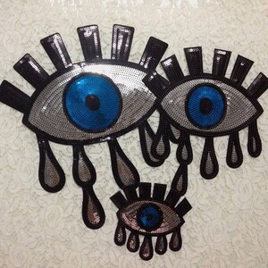 May include: Three silver sequin eye patches with blue pupils and black eyelashes. The larger eye patches have silver teardrop shapes hanging from the bottom.