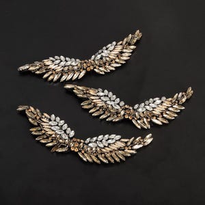 Puede incluir: Tres pares de alas de pedrería doradas y plateadas. Las alas están hechas de pequeños diamantes de imitación facetados y están dispuestas en forma de pluma.