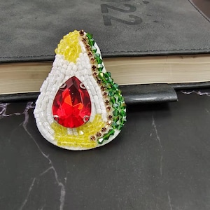 Puede incluir: Broche artesanal con forma de aguacate. Presenta una gran gema roja en forma de lágrima en el centro, rodeada de cuentas blancas. La pulpa del aguacate es blanca, con detalles de cuentas amarillas y verdes. Un accesorio único.