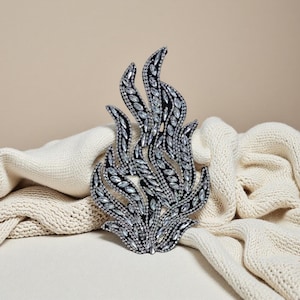 Puede incluir: Broche decorativo con forma de llama. El broche presenta una base negra con cristales transparentes y brillantes y detalles plateados. Se muestra sobre una tela de punto color crema.