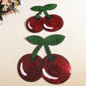 Könnte beinhalten: Zwei Paare roter und grüner Pailletten-Kirsch-Applikationen. Die Kirschen sind mit kleinen, runden Pailletten bestickt. Kirschen sind ein beliebtes Motiv für Kleidung und Accessoires.