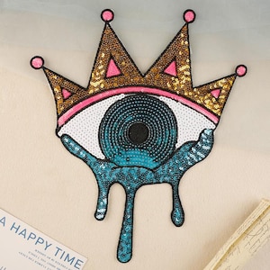 May include: A decorative sequined patch in the shape of an eye wearing a gold crown with pink details. The eye has a blue iris and teal teardrops. Suitable for embellishing clothing or accessories.