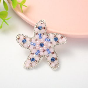 Può includere: Una stella marina di perline rosa e blu con strass sui bordi. La stella marina è su una superficie rosa.