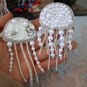 May include: Two white beaded jellyfish with pearl accents. The jellyfish are sewn with white beads and have dangling pearl strands.