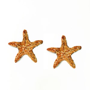 May include: Two decorative starfish with a shimmering gold sequin design. The starfish are arranged on a white background. These embellishments are ideal for craft projects or coastal-themed decor.