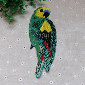 May include: A sequined parrot patch in shades of green, yellow, blue, and red. The bird's head is yellow with a blue beak and black accents. The body is green with black and silver details, and red accents on the legs. The patch is designed for decorative purposes.