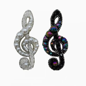 Puede incluir: Dos broches decorativos con forma de clave de sol. Uno es blanco con perlas de imitación y cristales transparentes. El otro es negro con cuentas iridiscentes. Ambos están sobre un fondo blanco. Estos accesorios de temática musical son ideales para añadir estilo.