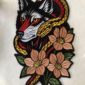 Puede incluir: Parche bordado con un lobo negro con detalles rojos y blancos, entrelazado con una serpiente amarilla y roja. El diseño incluye flores color melocotón con hojas verdes, todo sobre un fondo negro.