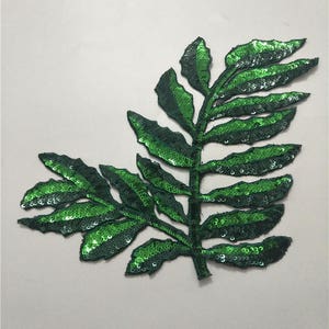Puede incluir: Aplicación decorativa de hoja con lentejuelas verdes. Las hojas están hechas de lentejuelas individuales, creando un efecto brillante. La aplicación está diseñada para ser cosida sobre tela, agregando un toque de brillo a la ropa o manualidades.