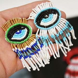 May include: A pair of beaded earrings with a blue evil eye design. The earrings have a fringe of white beads.